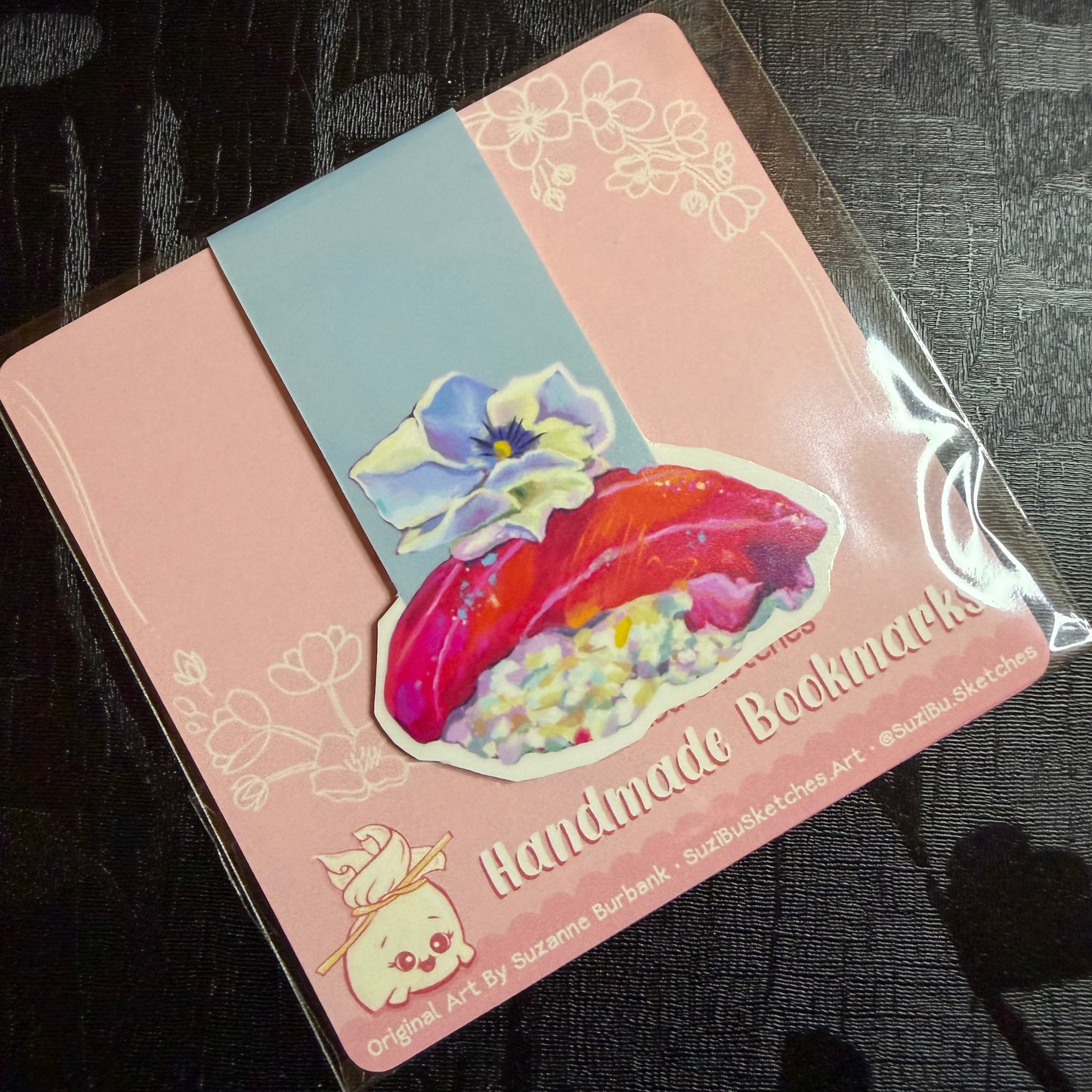 A packaged set of handmade bookmarks featuring a pink and light blue cover with a colorful illustration of a red tuna fish and flowers, sealed in clear plastic on a dark textured surface.