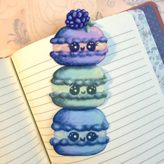 A cute digital illustration of three stacked macarons with cartoon eyes and smiles, placed on an open lined notebook. The top macaron is purple with a berry topper, the middle is mint green, and the bottom is blue.