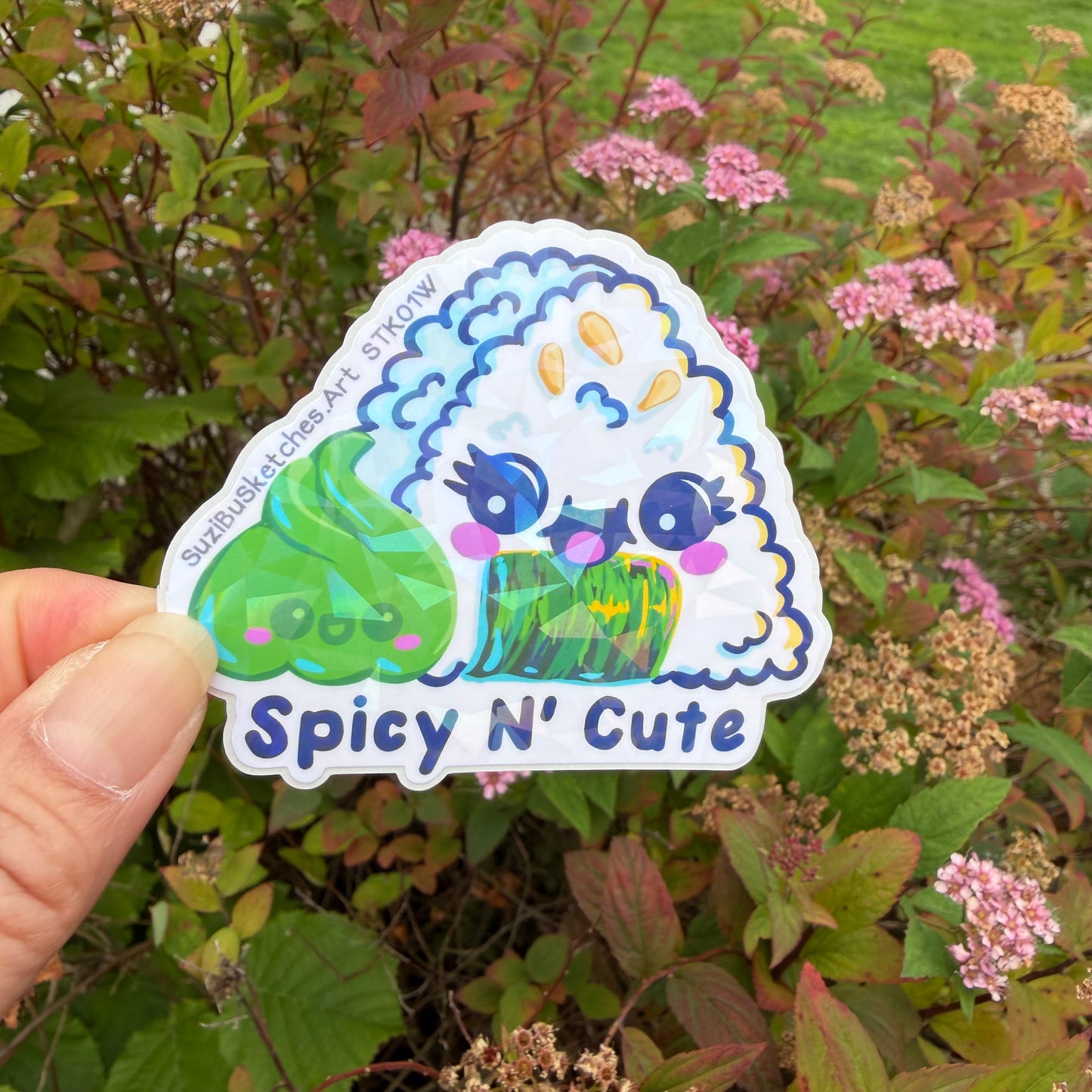 A hand holds a die-cut sticker featuring a cute cartoon rice onigiri character with the text 'Spicy N' Cute' and 'suzibusketches.art’ on a blurred garden background.