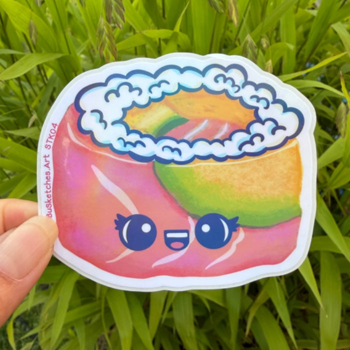 A hand holds a die-cut sticker featuring a cute, colorful cartoon sushi illustration with a smiling face, set against a background of green grass.