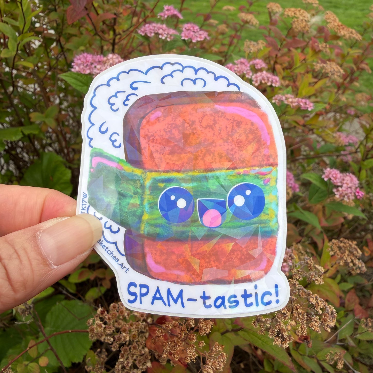 A hand holds a die-cut sticker featuring a cute, colorful cartoon spam Musubi with the text 'SPAM-tastic!' against a blurred garden background.