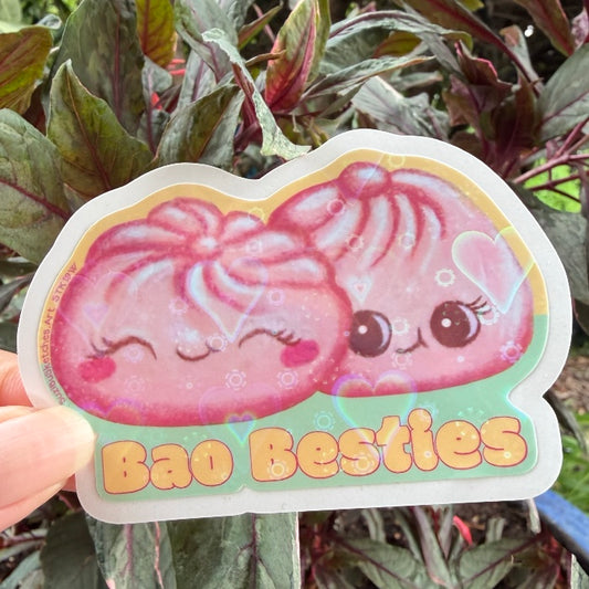 A hand holds a die-cut sticker featuring two cute pink bao buns with the text 'Bao Besties' on a green background, set against a backdrop of green and purple leafy plants.