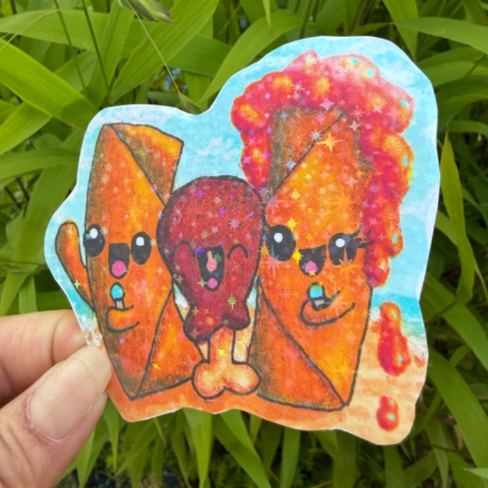 A hand holds a die-cut sticker featuring three cute, sparkly cartoon food characters against a background of green outdoor leaves.