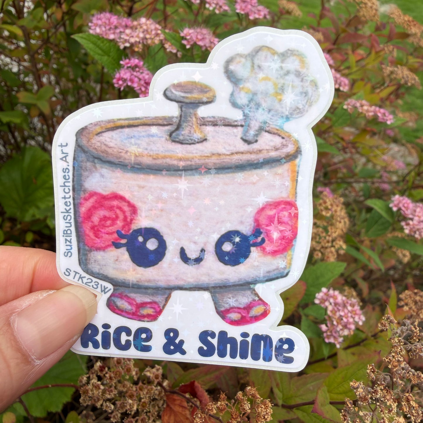 A hand holds a die-cut sticker featuring a cute cartoon kawaii rice character with pink flowers and the text 'Rice & Shine' against a blurred garden background of pink flowers and green leaves.