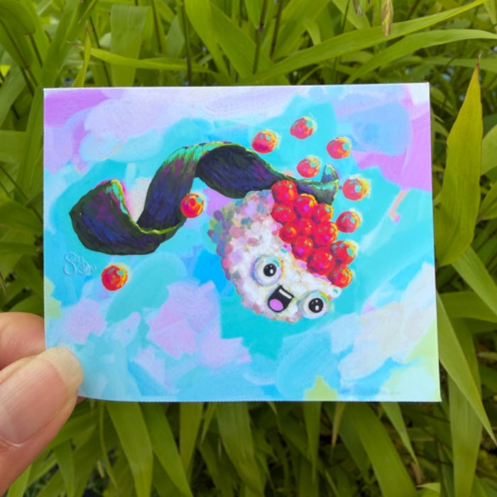A hand holds a small square art print or card featuring a whimsical cartoon creature with a dark green wavy body, red berry-like hair, and a light pink face against a pastel blue and purple background. The print is photographed outdoors in front of green grass.