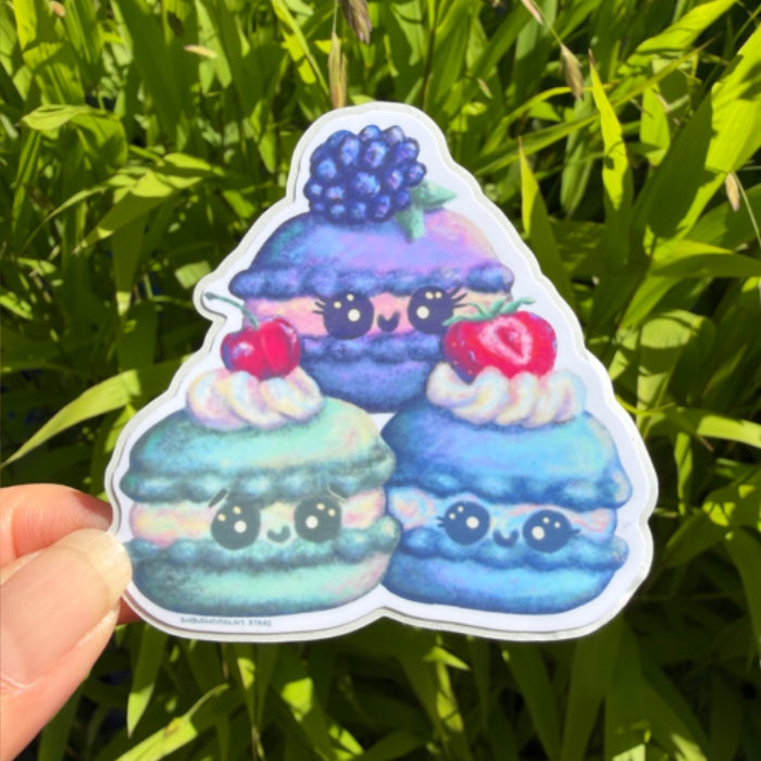 A hand holds a die-cut sticker featuring three cute, smiling macarons in shades of blue, purple, and green, topped with whipped cream and berries, set against a background of green grass.