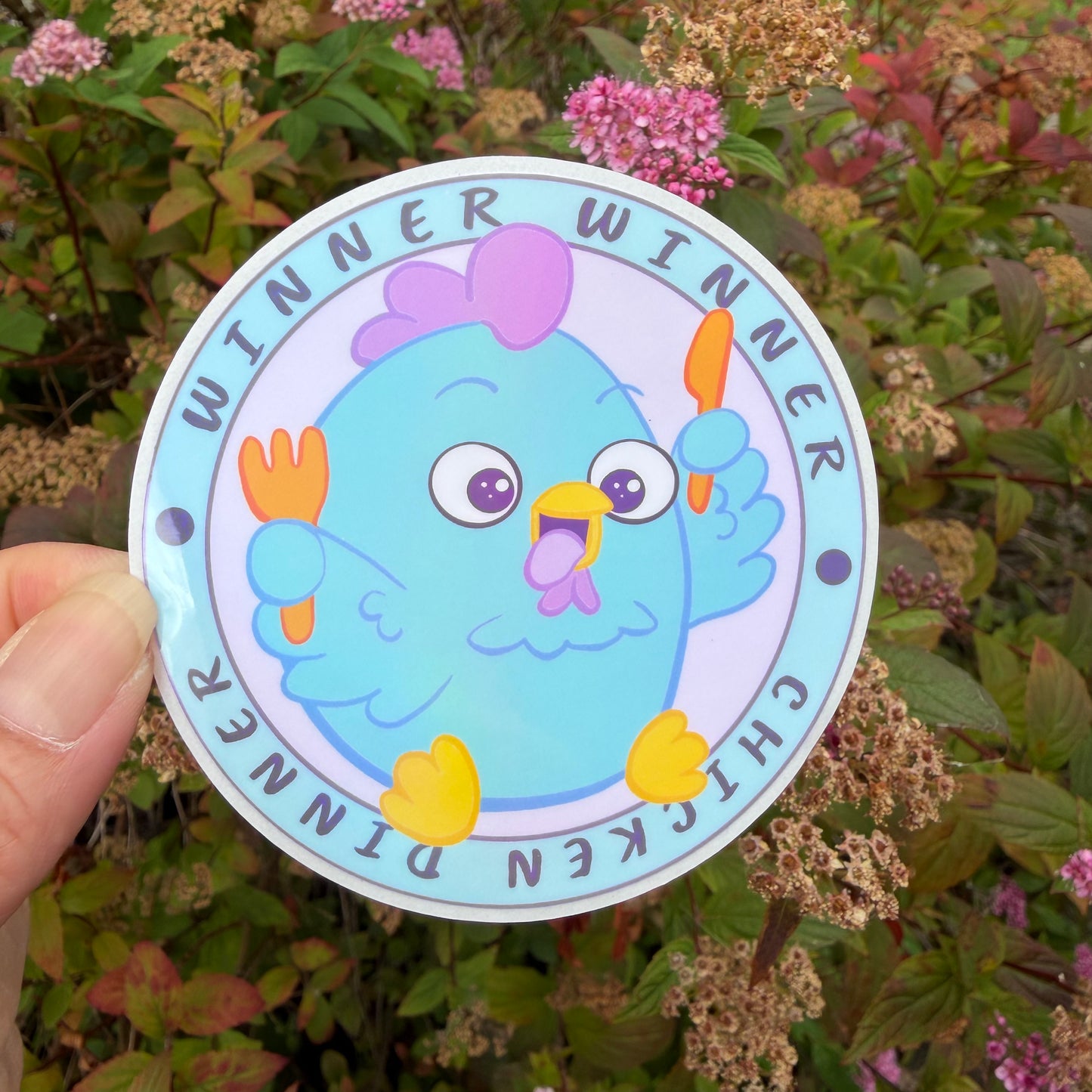 A hand holds a round sticker featuring a cartoon blue chicken holding orange forks, with the text 'WINNER WINNER CHICKEN DINNER' around the border, set against a background of green and pink flowers.