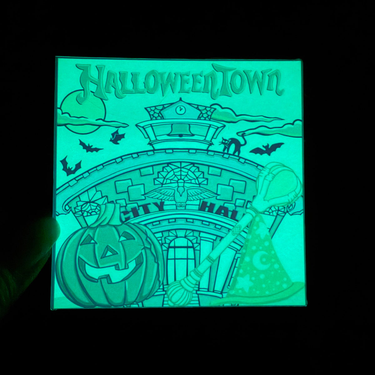 A square Halloween-themed light-up card or plaque glowing in the dark, featuring the words 'HALLOWEENTOWN' above an illustrated city hall, jack-o'-lantern, witch hat, bats, and moon. The product is photographed against a black background.