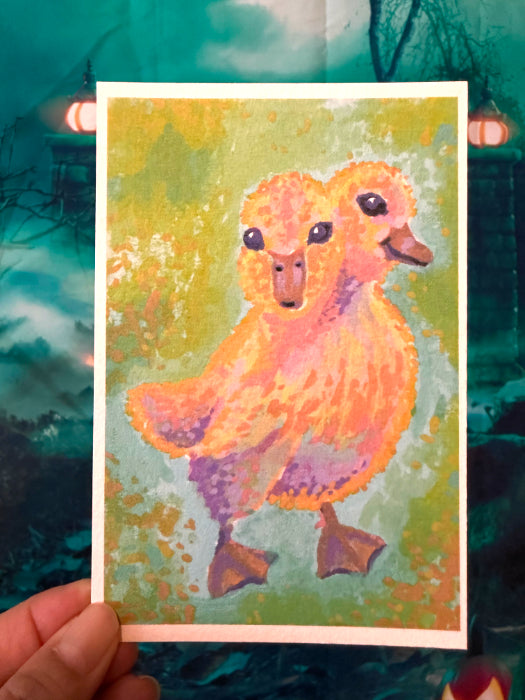 A hand holds a square art print or card featuring a painted 2-headed duckling in warm orange, pink, and purple tones against a green textured background. The print is photographed against a dark teal outdoor-like backdrop with blurred lights and branches.