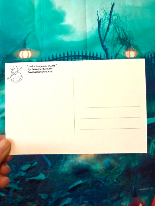 A hand holding a white postcard with the back side visible, featuring a small black sketch in the top left corner and the text 'Lucky Conjured Deck' by Suzanne Burbank. The background is a colorful illustrated scene with teal, blue, and black tones.