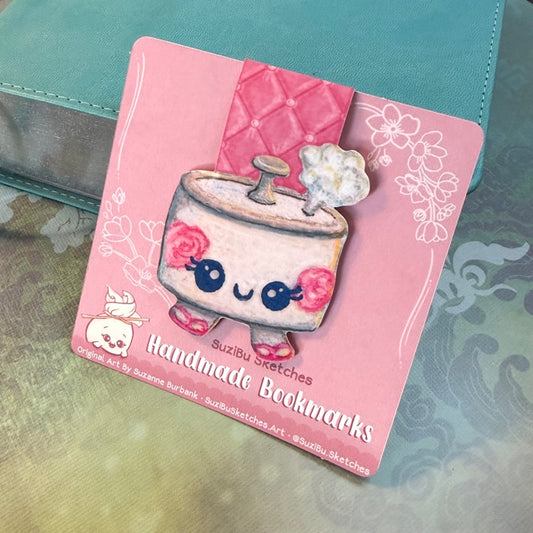 A pink card featuring a cute illustrated bookmark design of a kawaii rice cooker with the text 'Handmade Bookmarks' and 'SuziBu Sketches', placed on a teal leather surface.