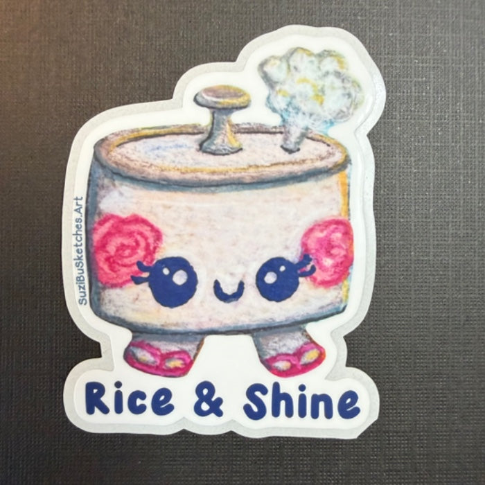 A die-cut sticker featuring a cute cartoon rice pot character with pink roses on its cheeks, blue eyes, and the text 'Rice & Shine' at the bottom, shown on a dark textured background.