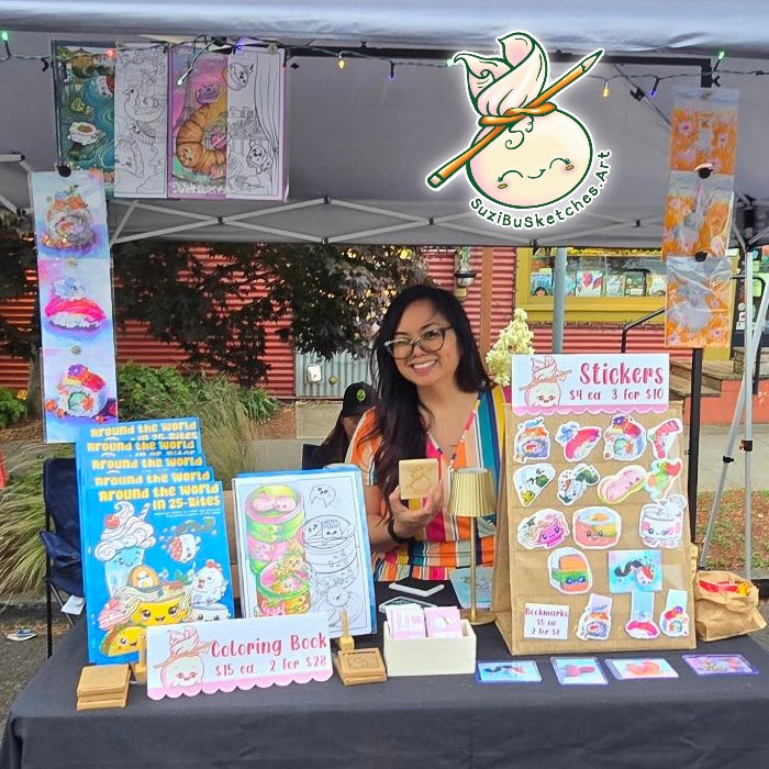 A smiling woman, Suzanne Burbank, stands behind a craft market booth displaying her art products, including stickers, a coloring book, and sketch prints hanging from a canopy. The booth has a logo overlay for 'SuziBuSketches.Art'.