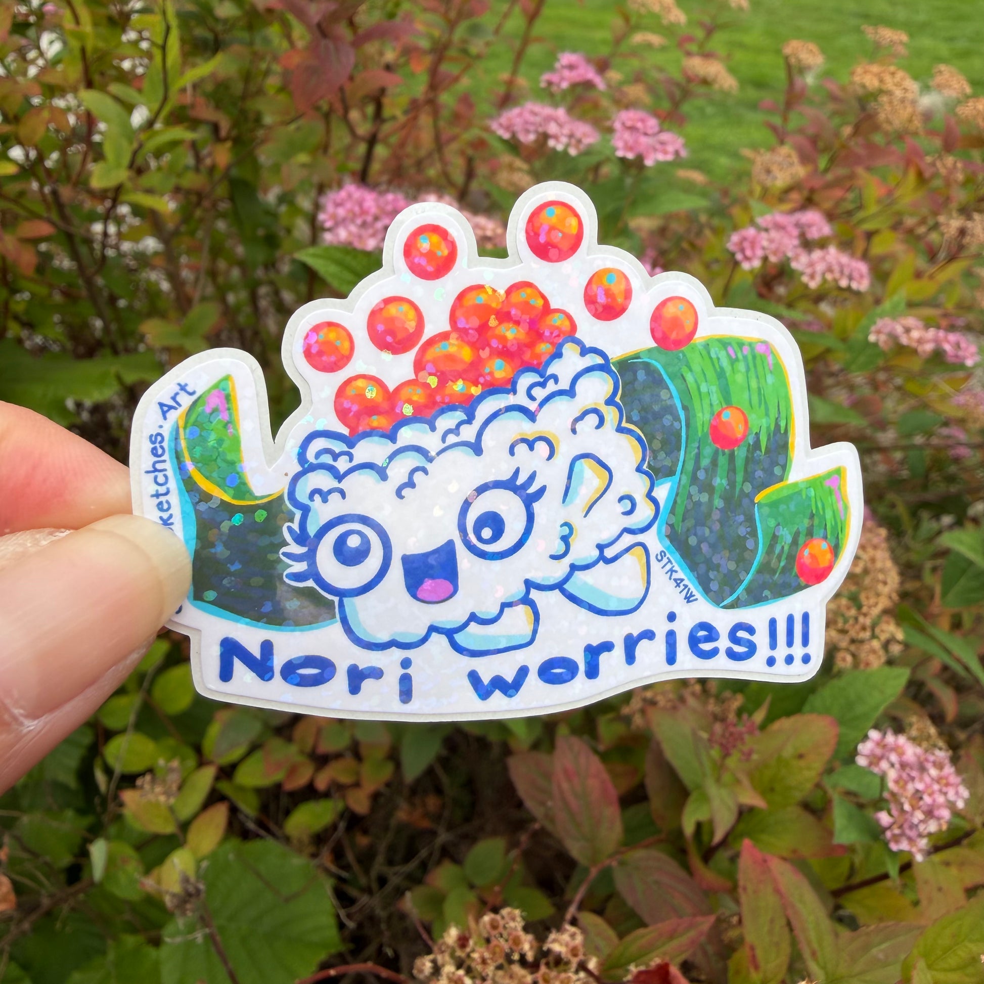 A hand holds a die-cut sticker featuring a cute kawaii cartoon character with the text 'Nori worries!!!' against a blurred garden background.