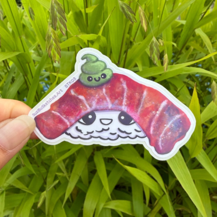 A hand holds a die-cut sticker shaped like a smiling sushi slice with a green washi ribbon, shown against a background of green grass.