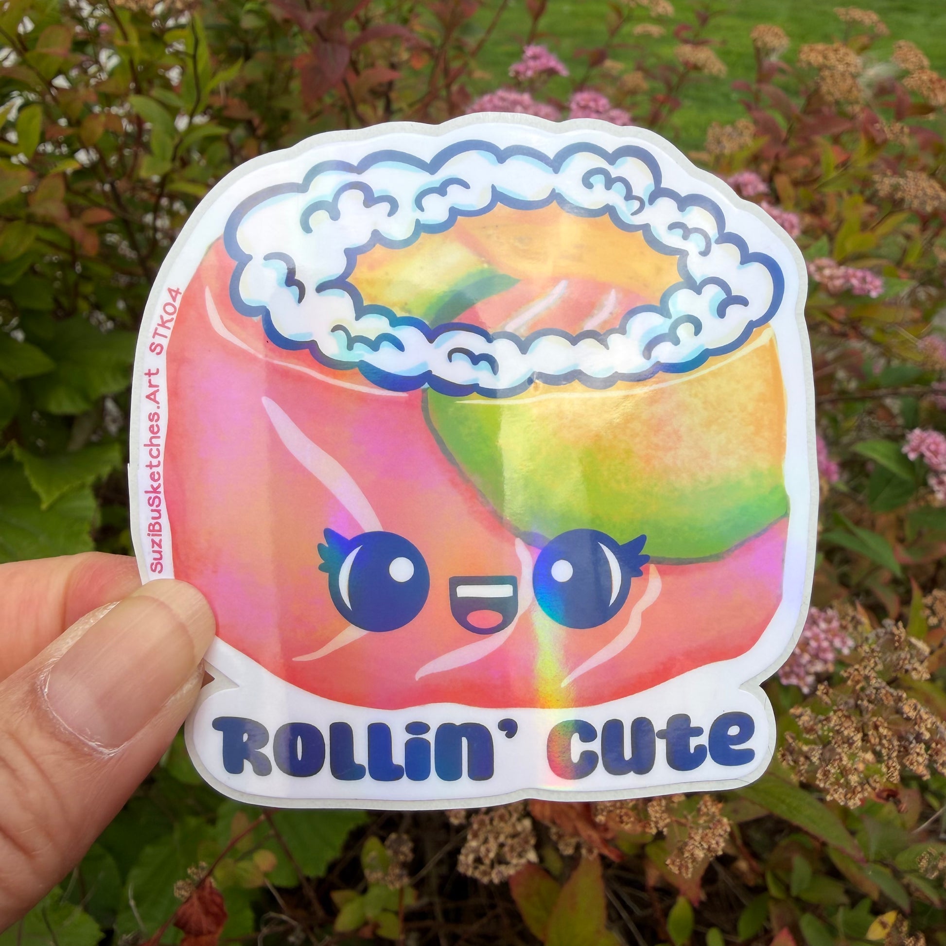 A hand holds a die-cut sticker featuring a colorful, cartoonish sushi salmon roll with a smiling face and the text 'ROLLIN' CUTE'. The sticker has a white border and is photographed outdoors against a blurred background of green foliage and pink flowers.