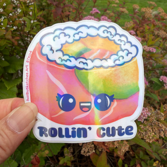 A hand holds a die-cut sticker featuring a colorful, cartoonish sushi salmon roll with a smiling face and the text 'ROLLIN' CUTE'. The sticker has a white border and is photographed outdoors against a blurred background of green foliage and pink flowers.