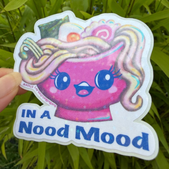 A hand holds a die-cut sticker featuring a pink bowl of ramen with a cute face and the text 'IN A Nood Mood'. The sticker is photographed outdoors against a blurred background of green leaves.