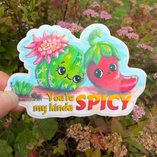 A hand holds a die-cut sticker featuring a green cactus and a red chili pepper with cartoon eyes and smiles, with the text 'You're my kinda SPICY'. The background is a blurred outdoor garden with green foliage and pink flowers.