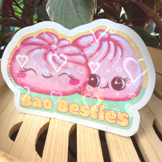 A die-cut sticker featuring a cute pink cartoon character with the text 'Bao Besties' in orange-yellow letters, displayed on a light wooden slatted surface with green leaves in the background.