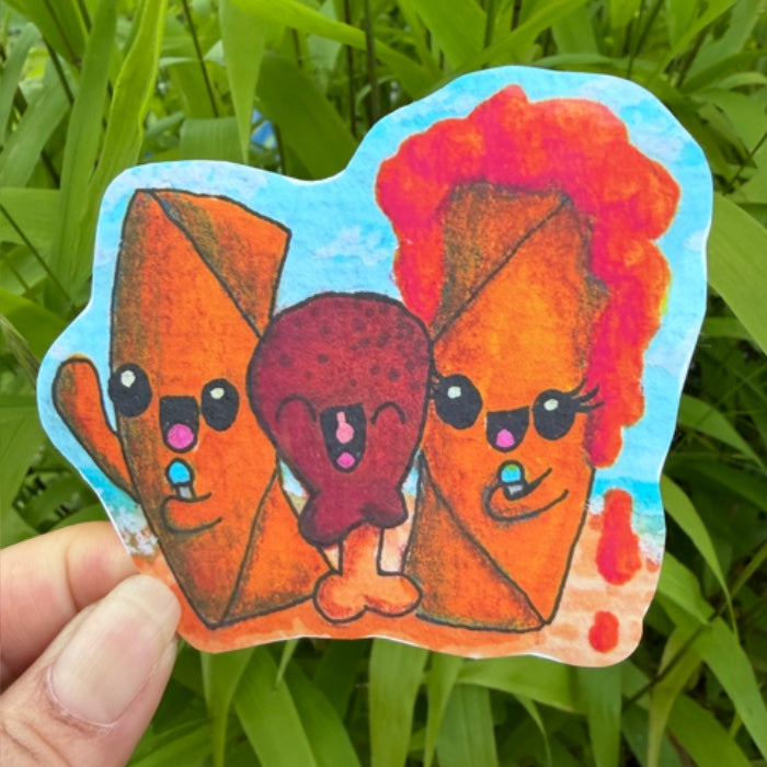 A hand holds a die-cut sticker featuring three cute, cartoonish food characters in orange and red tones against a light blue background, with green grass behind it.