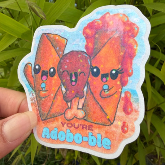 A hand holds a die-cut sticker featuring three cute cartoon food characters in orange and red tones against a light blue background, with the text 'YOU'RE Adobo-ble' at the bottom. Green leaves are visible in the background.