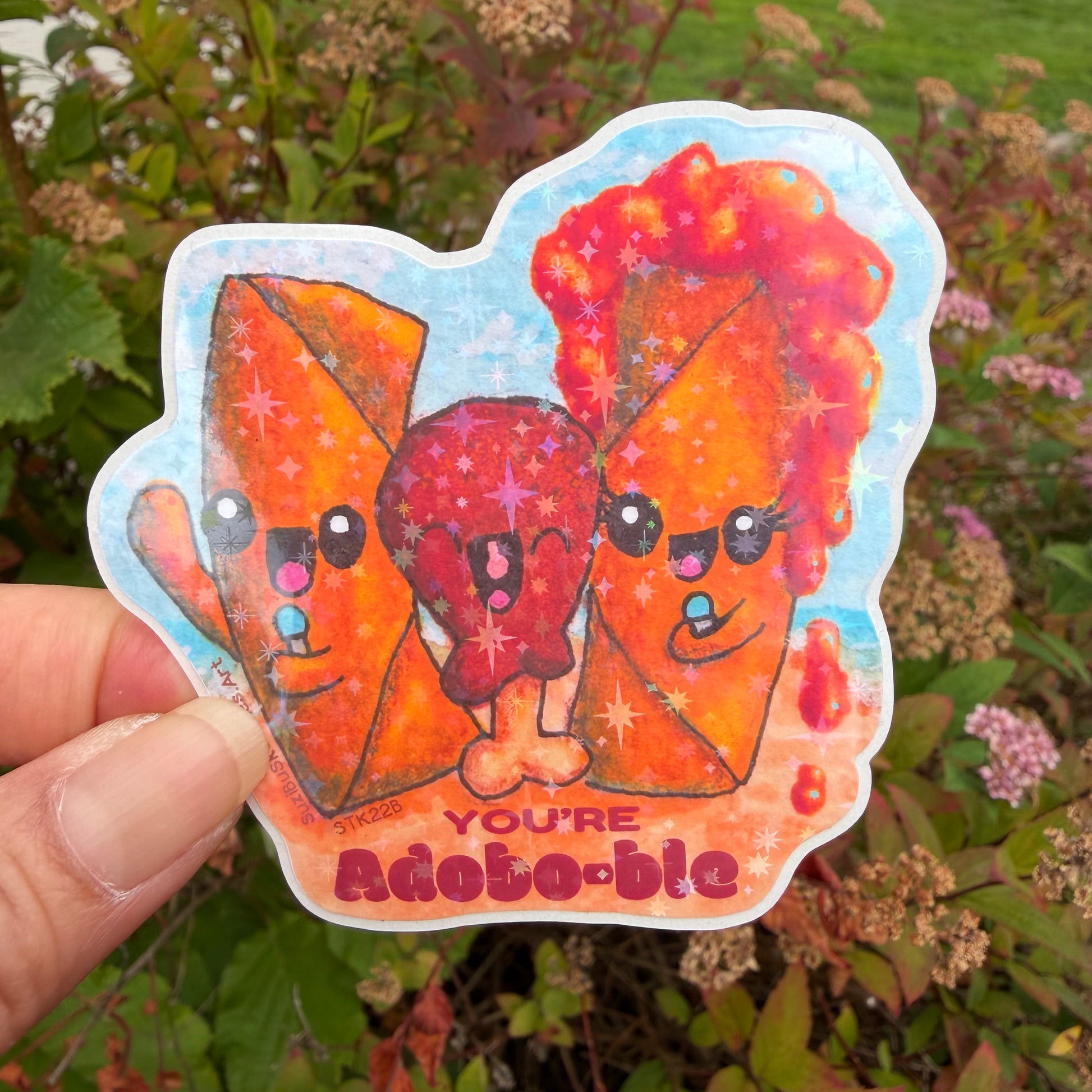 A hand holds a die-cut sticker featuring three cute, sparkly cartoon characters, lumpia and chicken adobo in orange and red tones against a blue background, with the text 'YOU'RE adobo-ble' at the bottom. The background is a blurred outdoor garden with green leaves and small pink flowers.