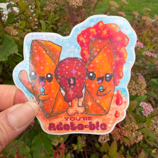 A hand holds a die-cut sticker featuring three cute, sparkly cartoon characters, lumpia and chicken adobo in orange and red tones against a blue background, with the text 'YOU'RE adobo-ble' at the bottom. The background is a blurred outdoor garden with green leaves and small pink flowers.