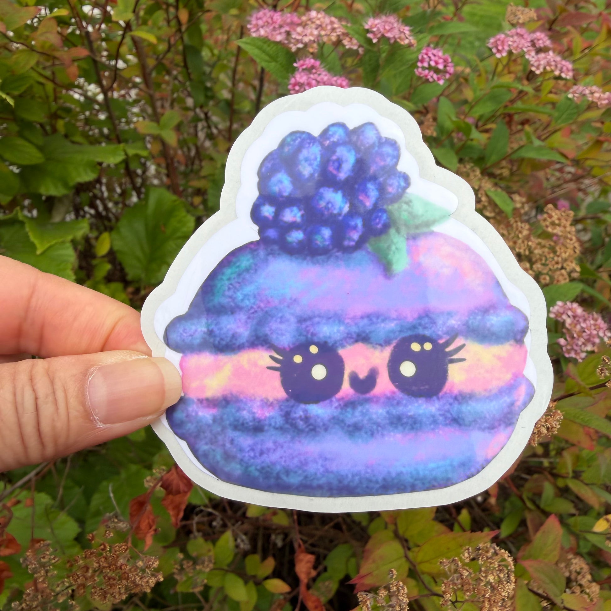A hand holds a die-cut sticker shaped like a cute macaron with a berry on top, photographed outdoors against green foliage and small pink flowers.