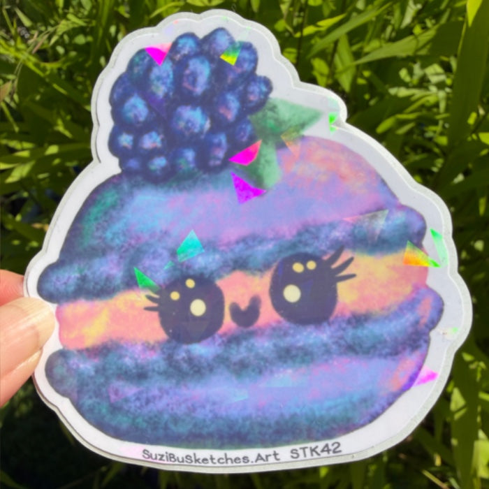 A die-cut sticker featuring a cute, kawaii-style illustrated character resembling a blueberry macaron with colorful pastel colors and a dark blue berry on top, held by a hand against a background of green foliage.