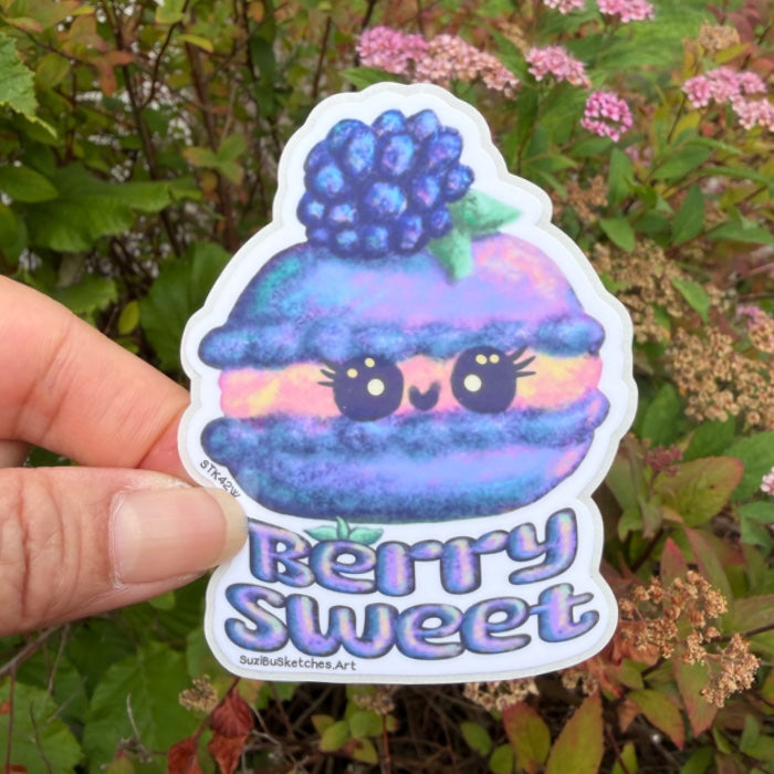 A hand holds a die-cut sticker featuring a cute macaron character topped with a berry and the words 'Berry Sweet' against a blurred garden background.
