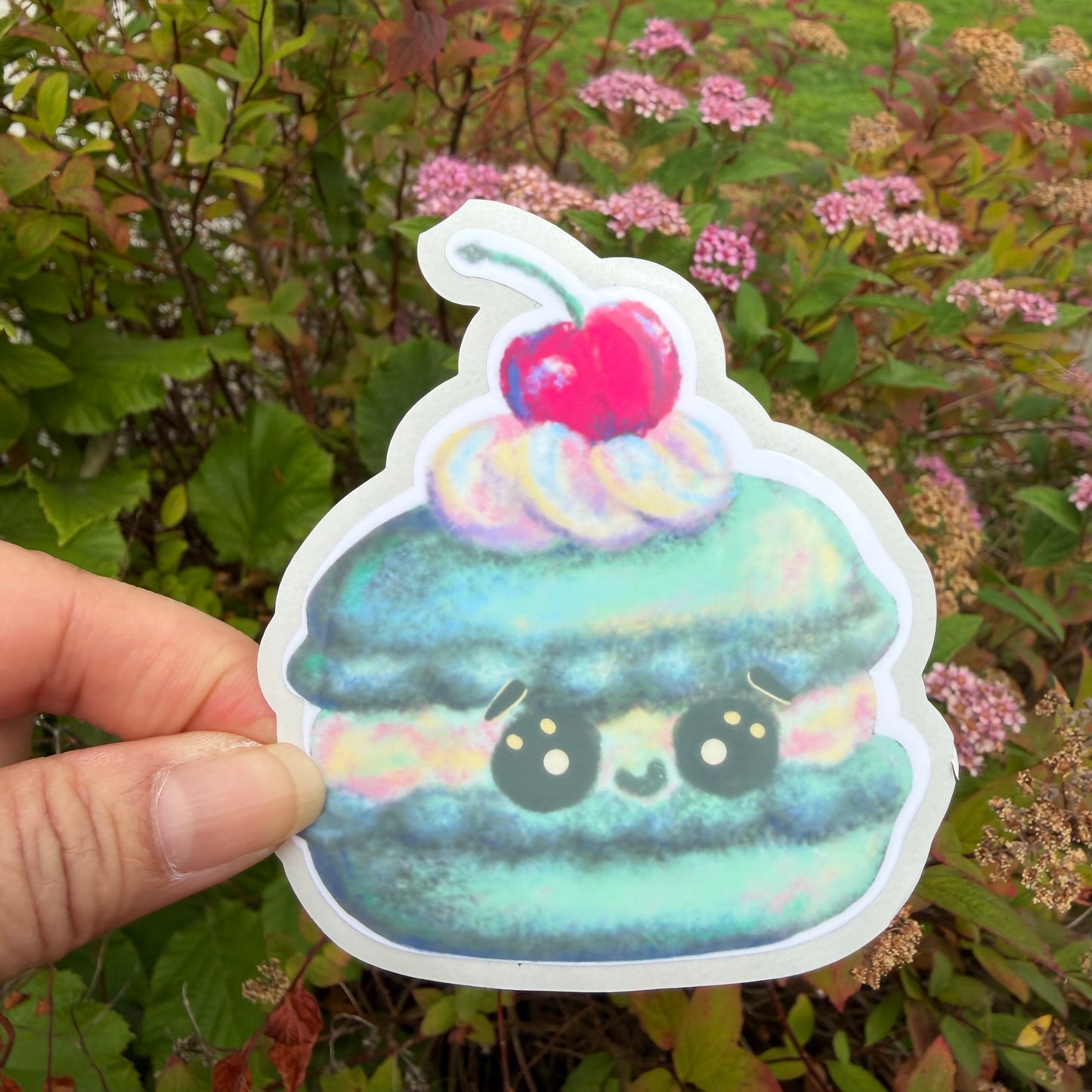 A hand holds a die-cut sticker of a cute, smiling kawaii macaron with a cherry on top, set against a background of green foliage and pink flowers.