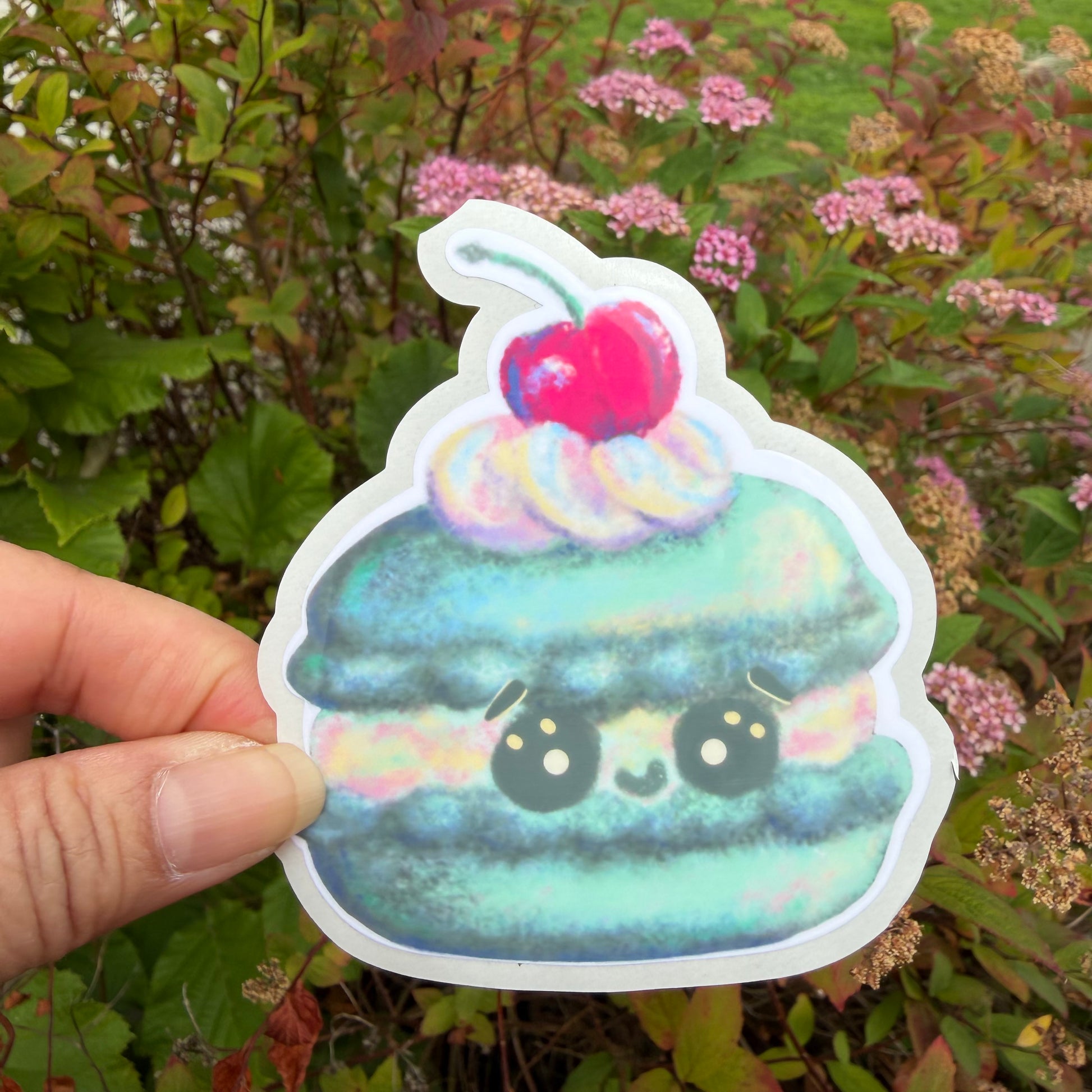 A hand holds a die-cut sticker of a cute, smiling kawaii macaron with a cherry on top, set against a background of green foliage and pink flowers.