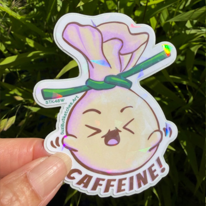 A die-cut sticker featuring a cartoon dumpling character with the word 'CAFFEINE!' at the bottom, held by a hand against a background of green foliage. The sticker has holographic/iridescent accents.
